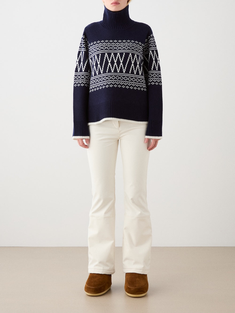 A person stands against a plain background wearing the We Norwegians Setesdal Pullover women sweater, featuring navy blue with white geometric patterns, paired with off-white pants and brown suede boots for a cozy winter look.