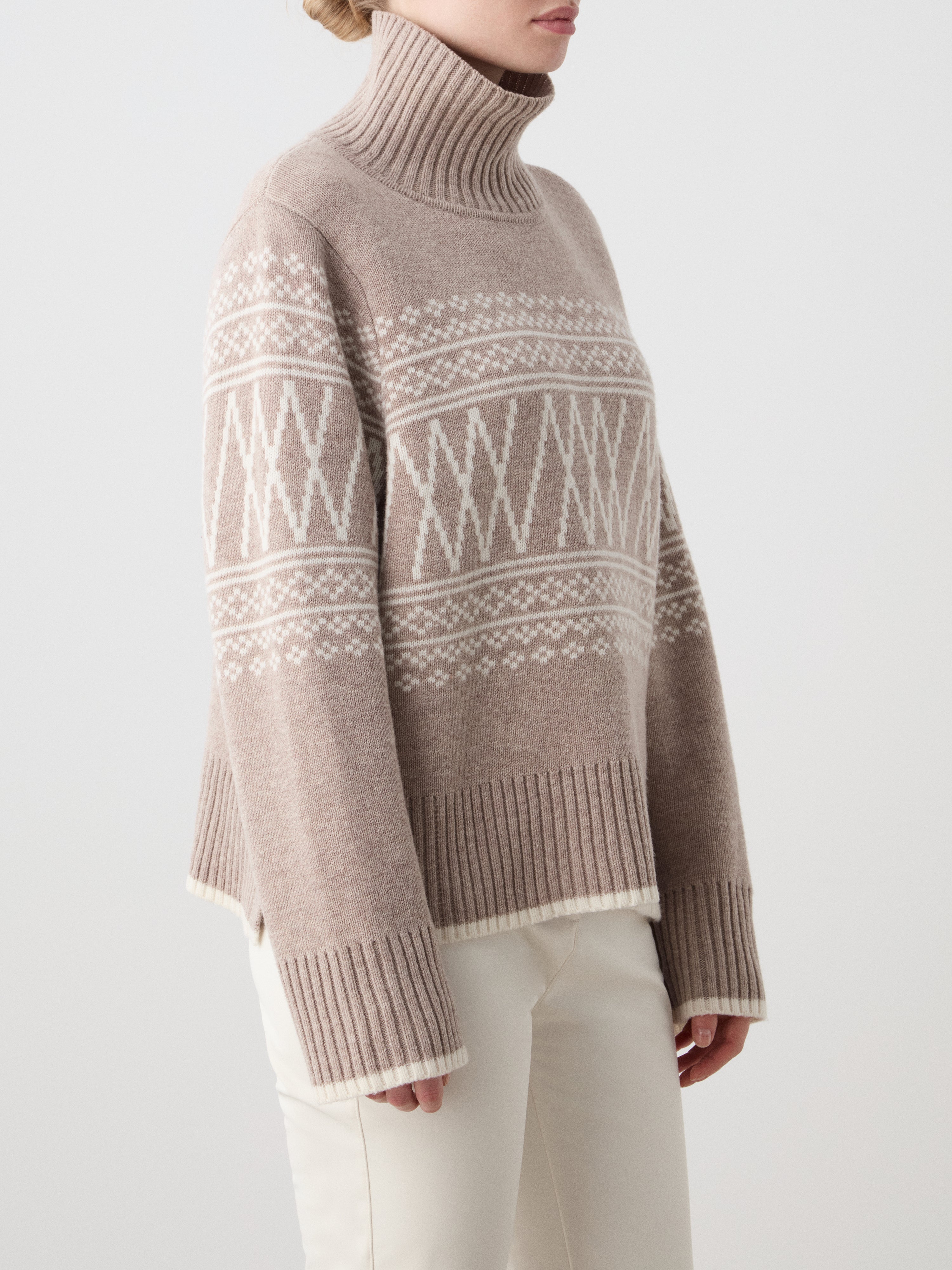 A woman wears the We Norwegians Setesdal Pullover in beige with white geometric patterns and cream-colored pants, standing against a plain light background.