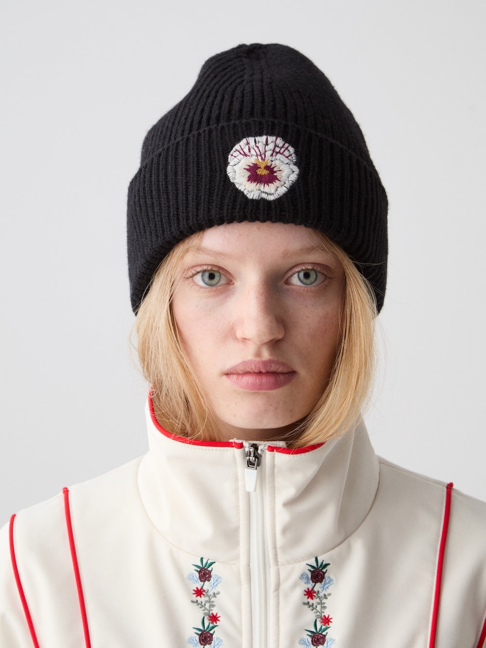A woman with light skin and straight blonde hair wears the We Norwegians WN x Markarian Hat Women, a limited edition black knit beanie with an embroidered patch, and a white floral embroidered jacket with red piping against a plain light background.