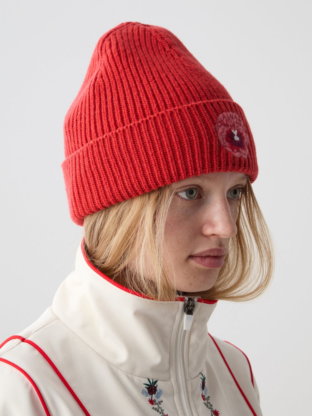 A woman wears the WN x Markarian Hat Women by We Norwegians, a limited edition red knit beanie with a small round patch, paired with a white zip-up jacket embroidered with small flowers against a plain background.