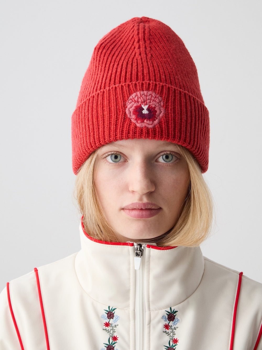 A blonde woman wears the We Norwegians WN x Markarian Hat Women, a limited edition red knit beanie with a circular patch, paired with a white floral embroidered zip-up top, standing against a plain light background.