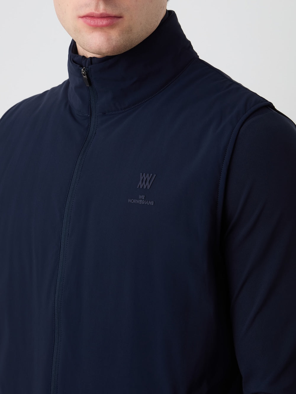 A man wears the We Norwegians Albatross Vest Men in navy—a weather-resistant golf vest with a subtle logo and "WE NORWEGIANS" on the left chest. Only his lower face and upper torso are shown.