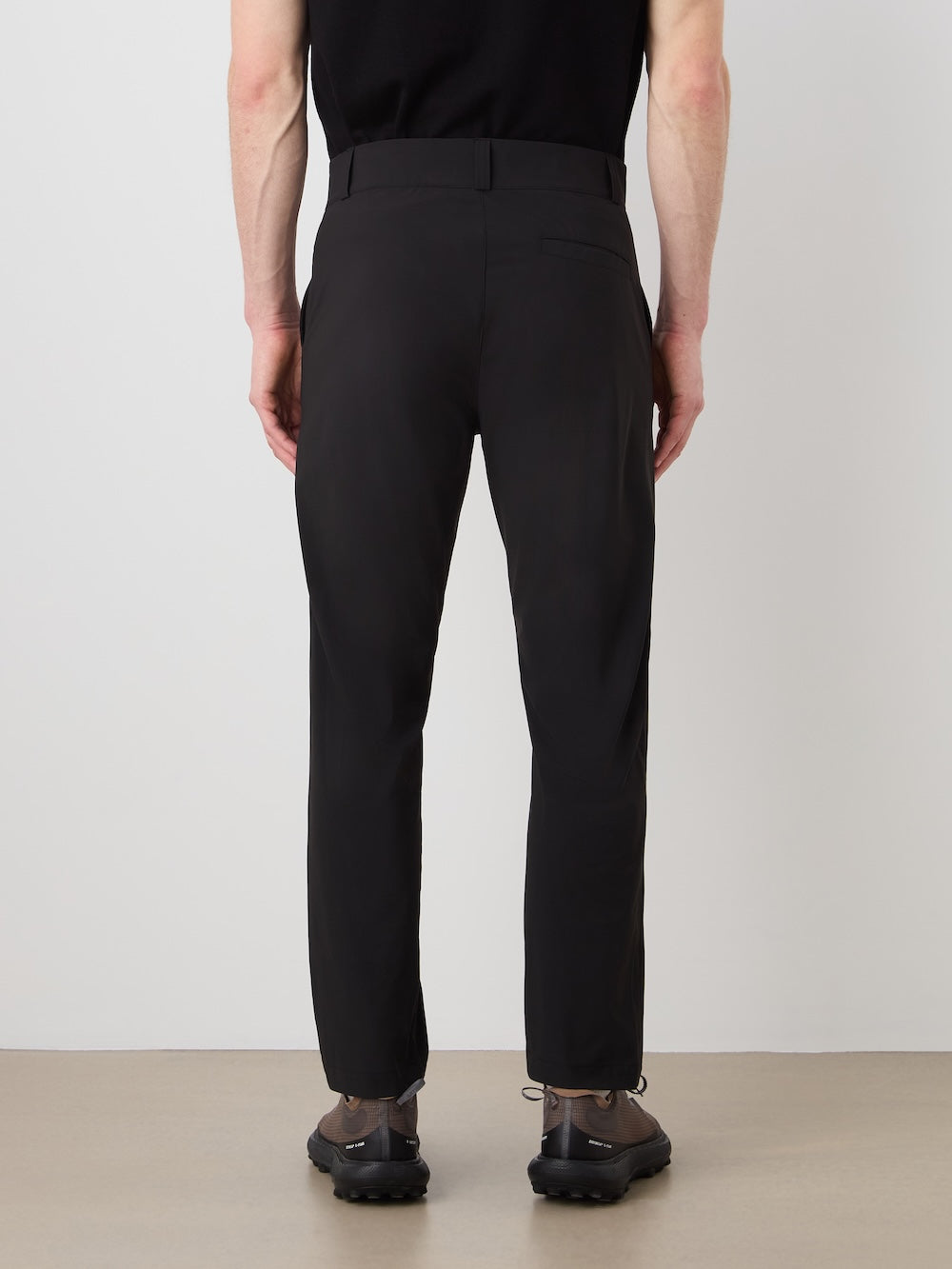 A person is seen from behind indoors wearing We Norwegians Albatross Pant Men in black—crafted with 4-way stretch, breathable performance fabric—paired with a black shirt and dark shoes, standing against a plain light-colored wall.
