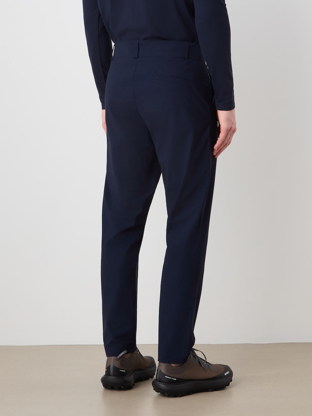 A person stands indoors facing away, wearing We Norwegians' Albatross Pant Men in navy blue with a matching long-sleeve top and dark brown thick-soled shoes. The scene shows a plain white wall and light brown floor.