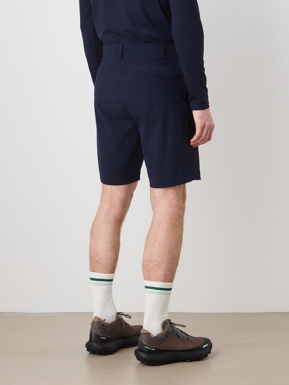 A person stands facing away, wearing a navy long-sleeve shirt and We Norwegians Albatross Shorts Men made from breathable stretch fabric, paired with white socks with green stripes and brown sneakers against a light wall and floor.