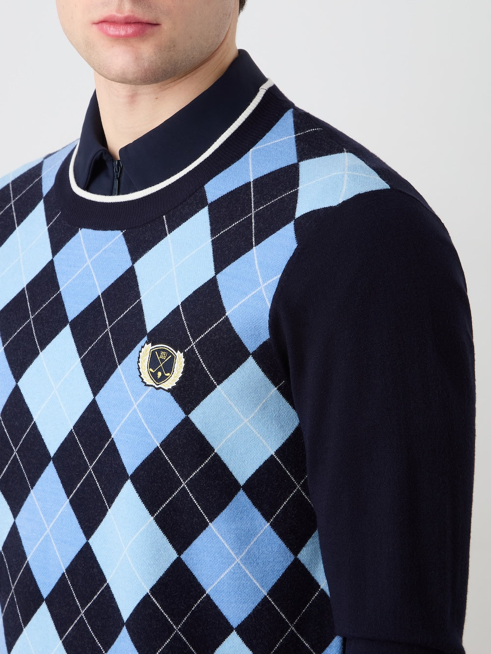 A man wears the We Norwegians Fairway Sweater Men—a blue and navy argyle-knit, gold and black-crested sweater—over a navy polo. Only his lower face and upper torso are visible.
