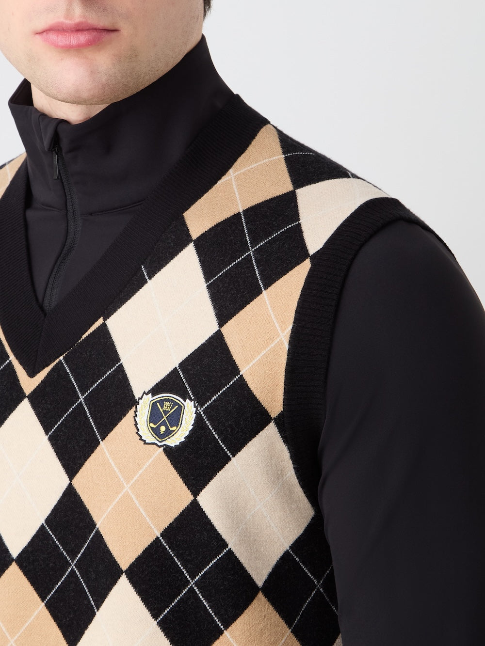 A man models the We Norwegians Fairway Vest Men, a beige, black, and white argyle knit golf vest with a crest patch on the chest, layered over a black zip-up top. Only his lower face is visible.