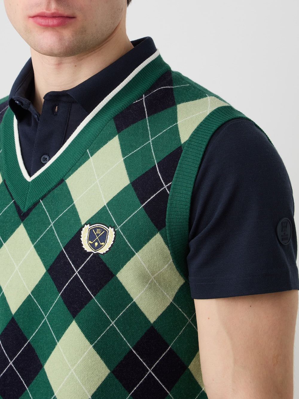 A person is seen from the nose down wearing the We Norwegians Fairway Vest Men, an argyle knit sleeveless sweater in green, black, and white with a small chest crest, over a dark navy polo shirt.