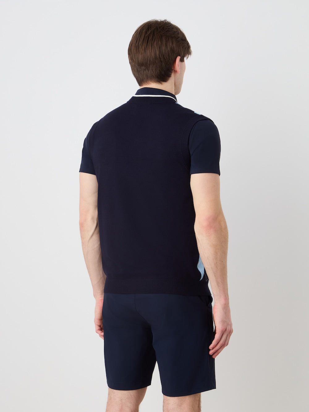 A man with short brown hair, wearing the We Norwegians Fairway Vest Men in navy and matching shorts, stands facing away from the camera against a plain light background.