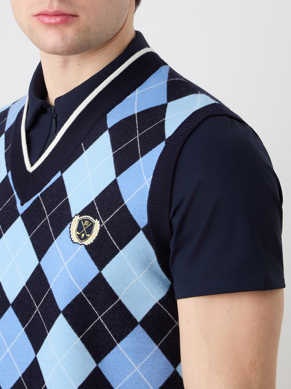 A man wears the We Norwegians Fairway Vest Men, a blue and black argyle knit golf vest with white trim and an embroidered crest, layered over a navy short-sleeve shirt. The image captures his upper body and part of his face.