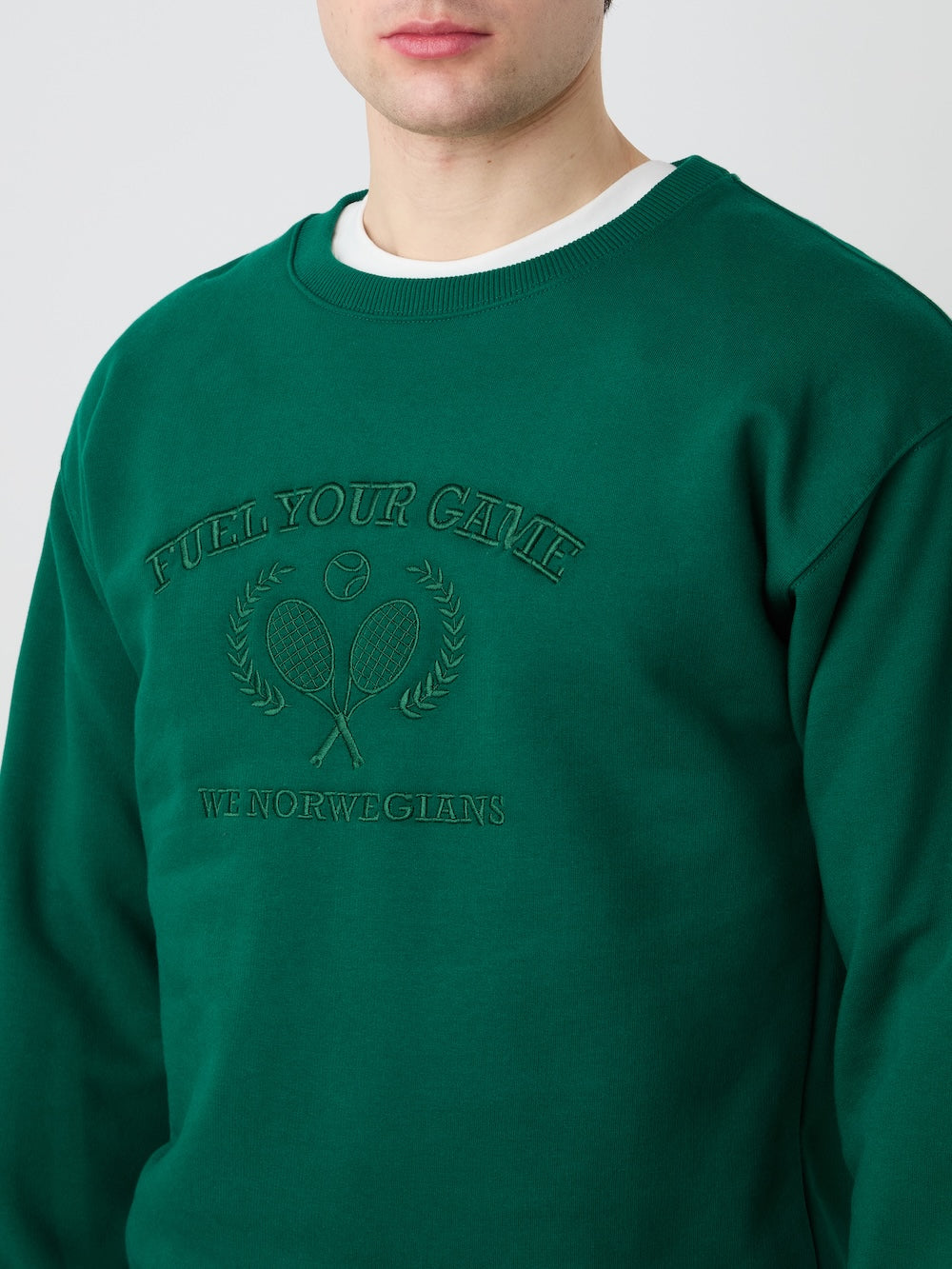 A man wears the We Norwegians Bounce Sweater Men, a green vintage tennis-style velour crewneck with "FUEL YOUR GAME" above crossed tennis rackets and ball, and "WE NORWEGIANS" below; only his lower face is visible.