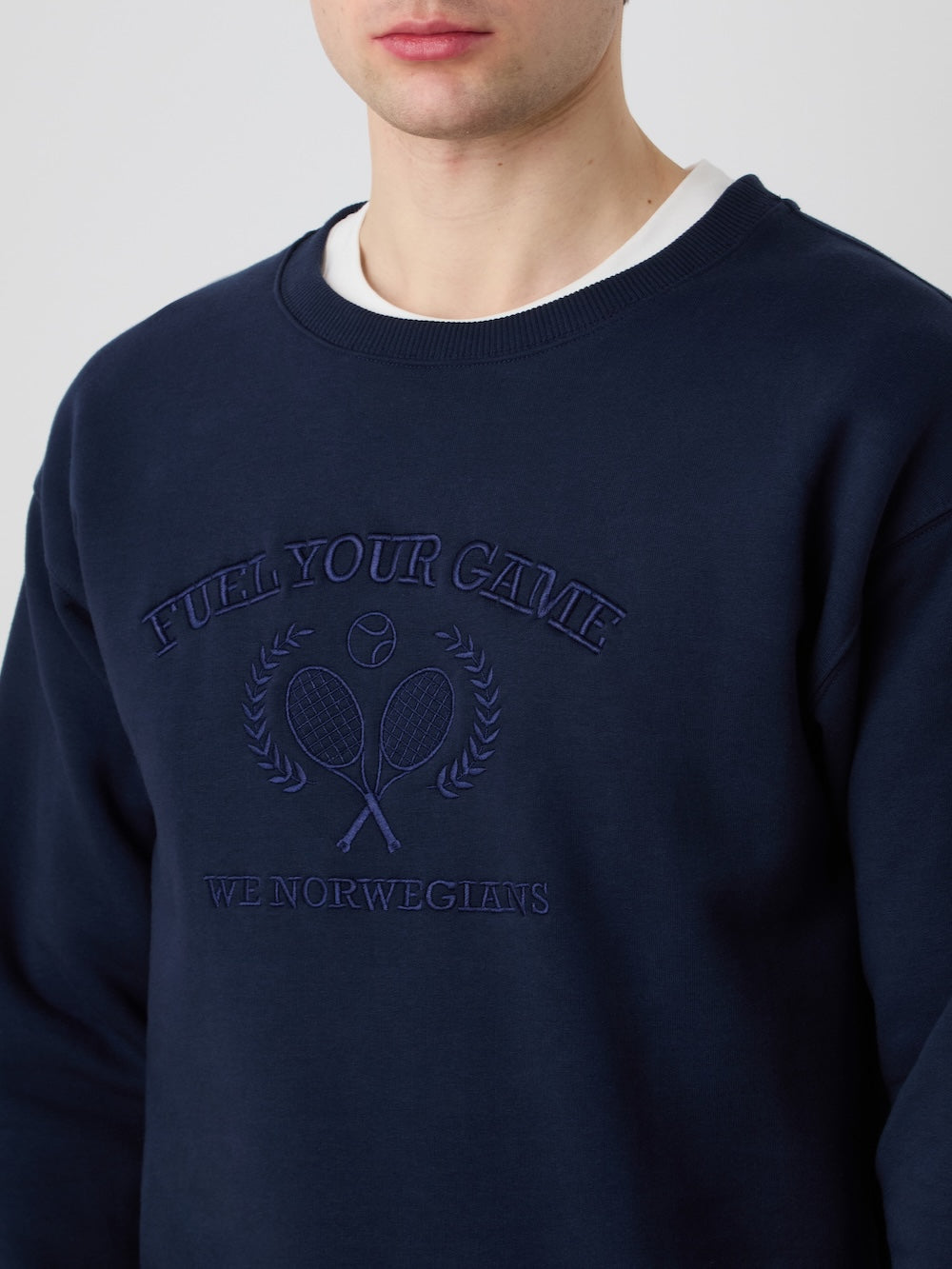 A person wears the We Norwegians Bounce Sweater Men in navy blue, featuring "FUEL YOUR GAME" and "WE NORWEGIANS" embroidered above two crossed tennis rackets and a ball, channeling a vintage tennis style. The face is partially visible.