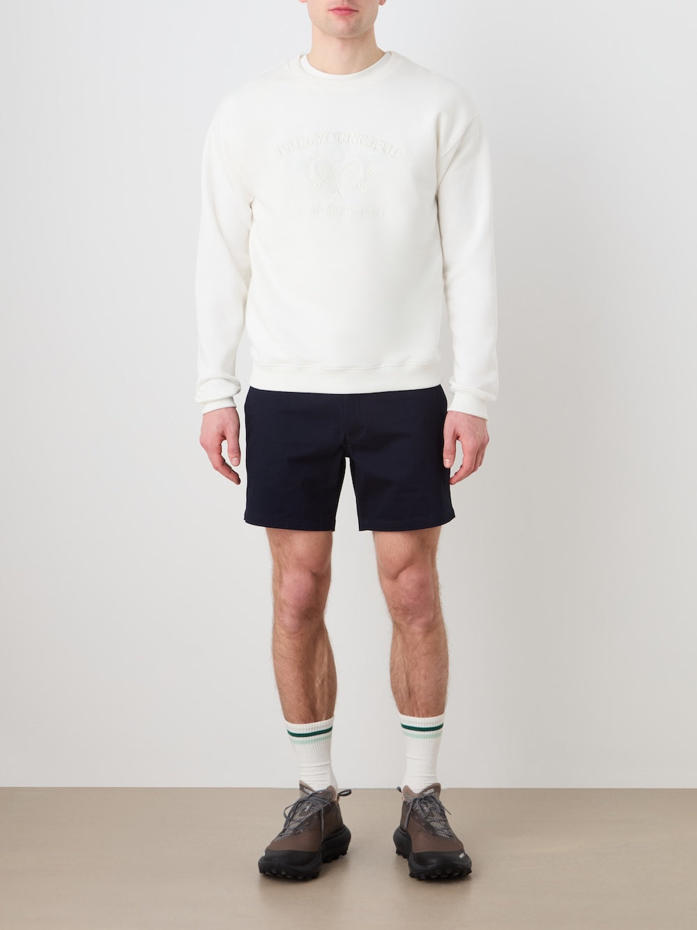 A man stands against a white wall wearing the We Norwegians Bounce Sweater Men, navy shorts, gray lace-up shoes, and white socks with green stripes for vintage tennis flair. His hands are by his sides; head slightly out of frame.