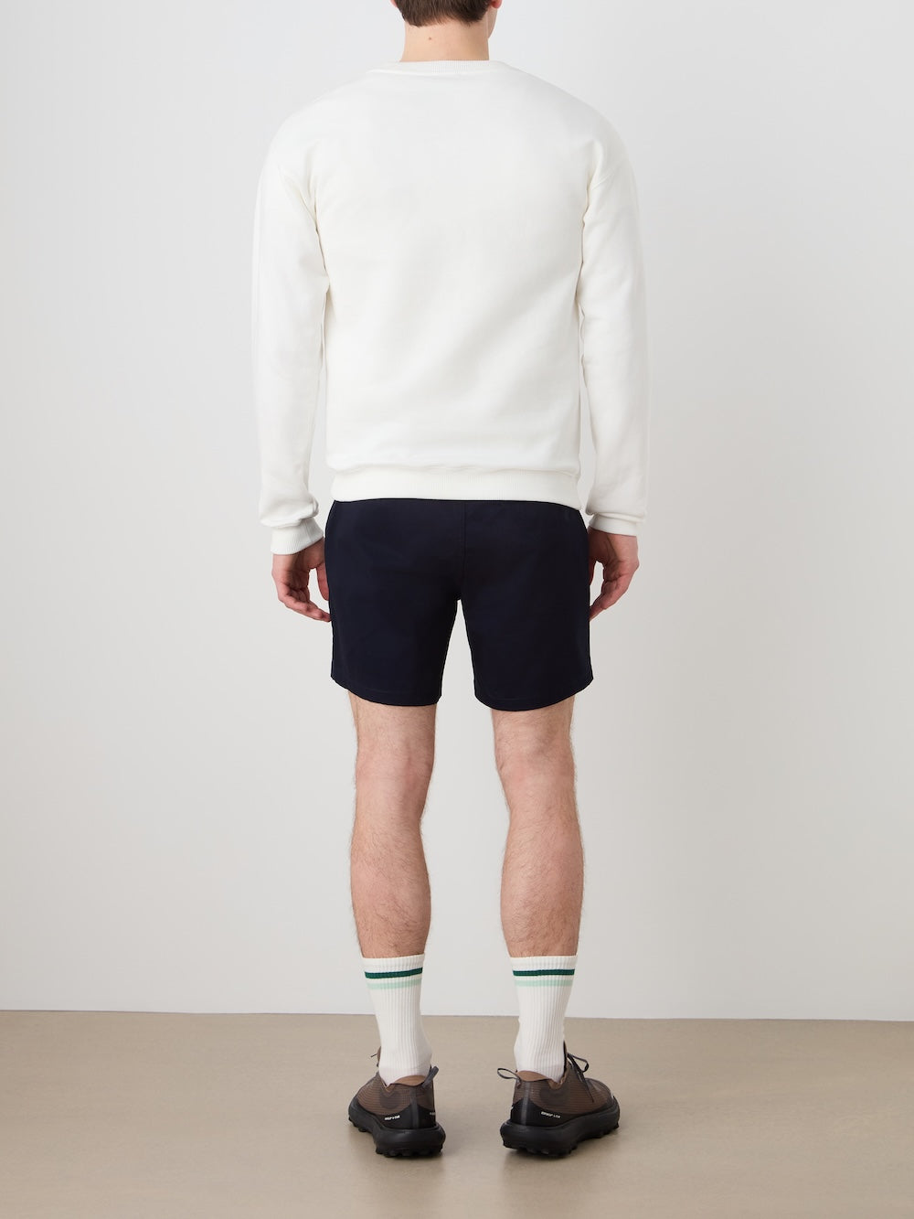 A man faces away from the camera, wearing the We Norwegians Bounce Sweater Men, navy shorts, white socks with green stripes, and dark shoes, channeling a vintage tennis style against a plain background.