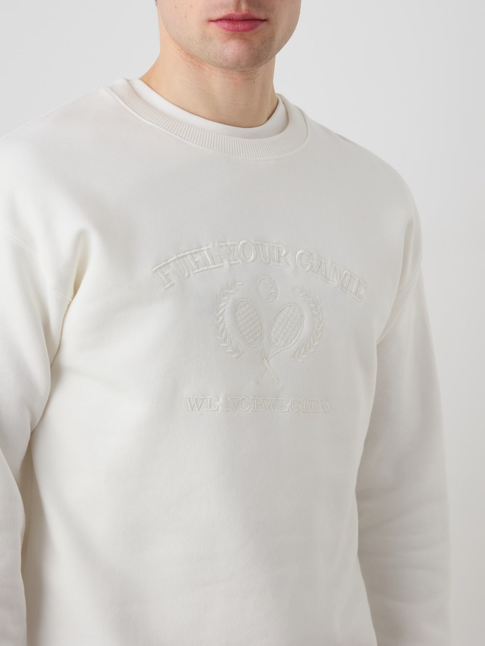 A man wears the We Norwegians Bounce Sweater Men in off-white velour, embossed with two tennis rackets, laurel leaves, and "PULL YOUR CANOE," showcasing a vintage tennis-inspired look.