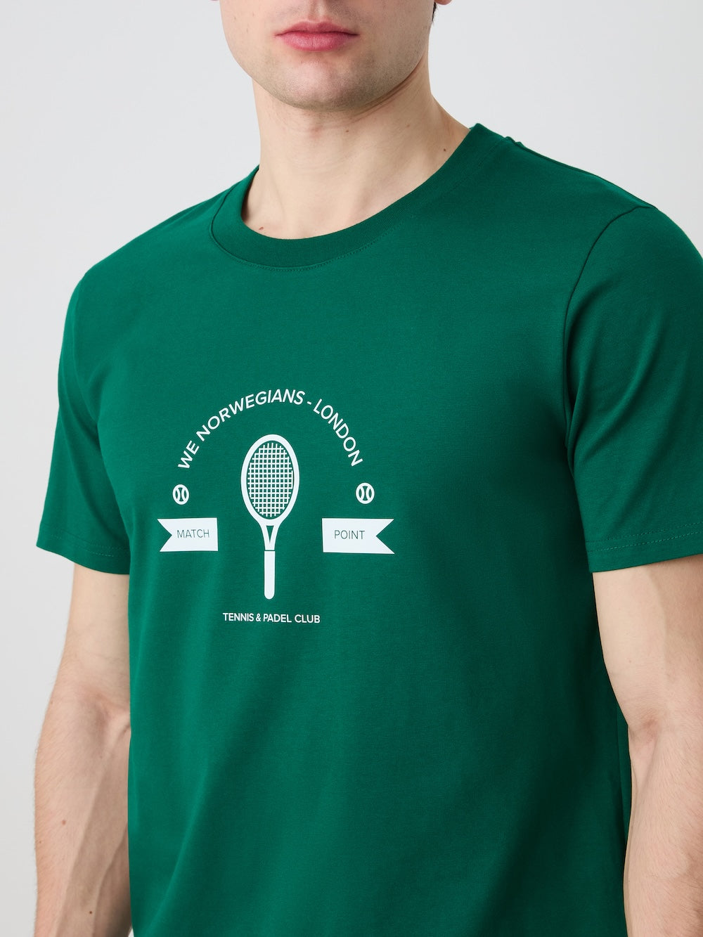 A man wears the We Norwegians Love Tee Men in green, showcasing a white racket graphic and “WE NORWEGIANS - LONDON,” “MATCH POINT,” and “TENNIS & PADEL CLUB.” The vintage style recalls retro tennis posters. Face is partly out of frame.