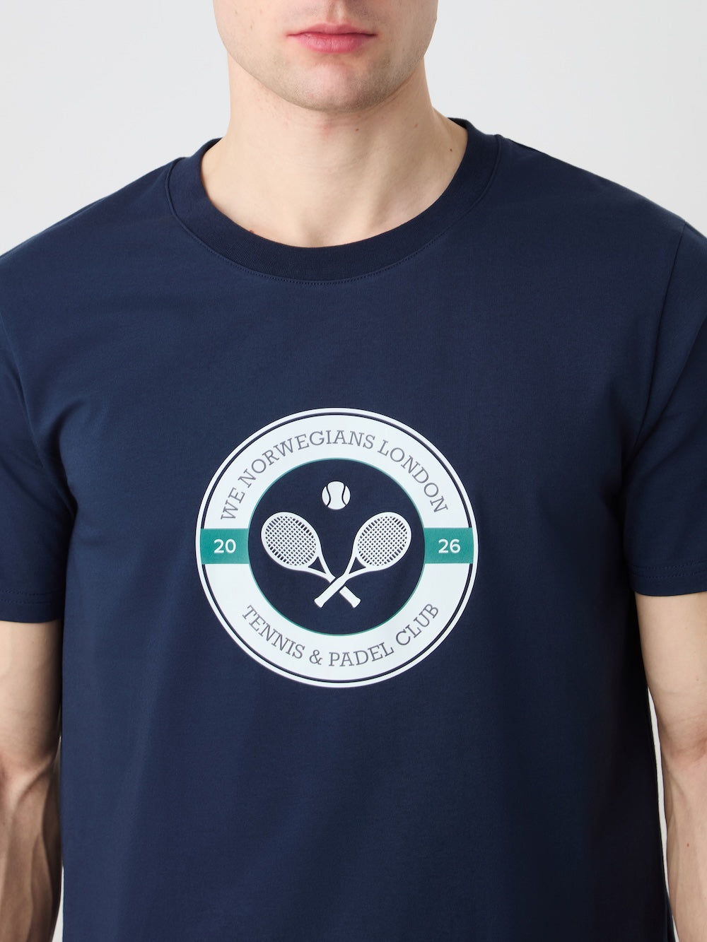 A man wears the Love Tee Men by We Norwegians, a navy cotton T-shirt with a circular logo of crossed rackets, a tennis ball, and "The Norwegians London Tennis & Padel Club 2026," inspired by retro tennis posters.