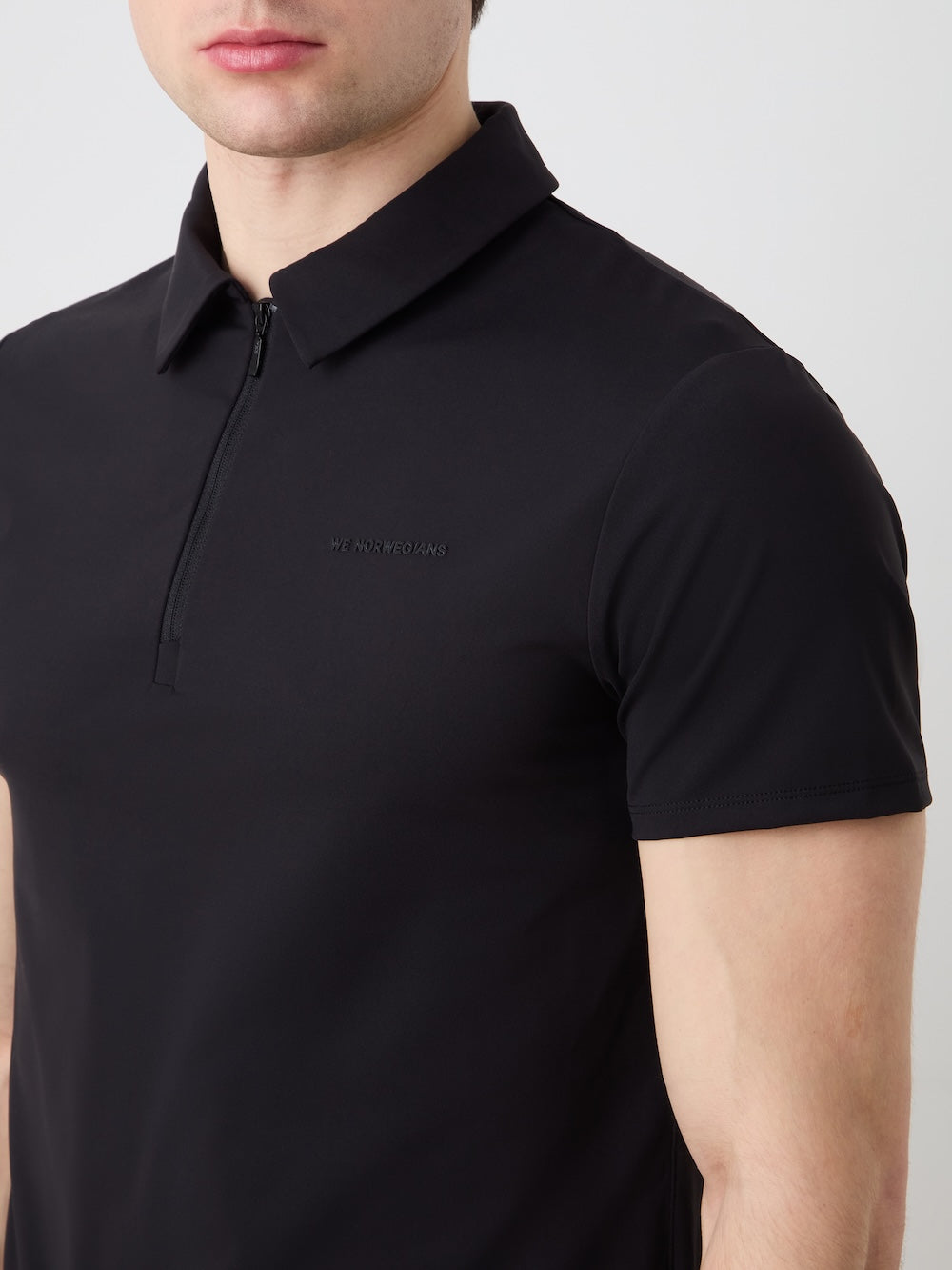 A man wears the We Norwegians Honor Zipped Polo Men—a black, high-performance polo with a chest logo and zipper—against a plain light background. The shirt delivers comfort and style, with his face partially out of frame.