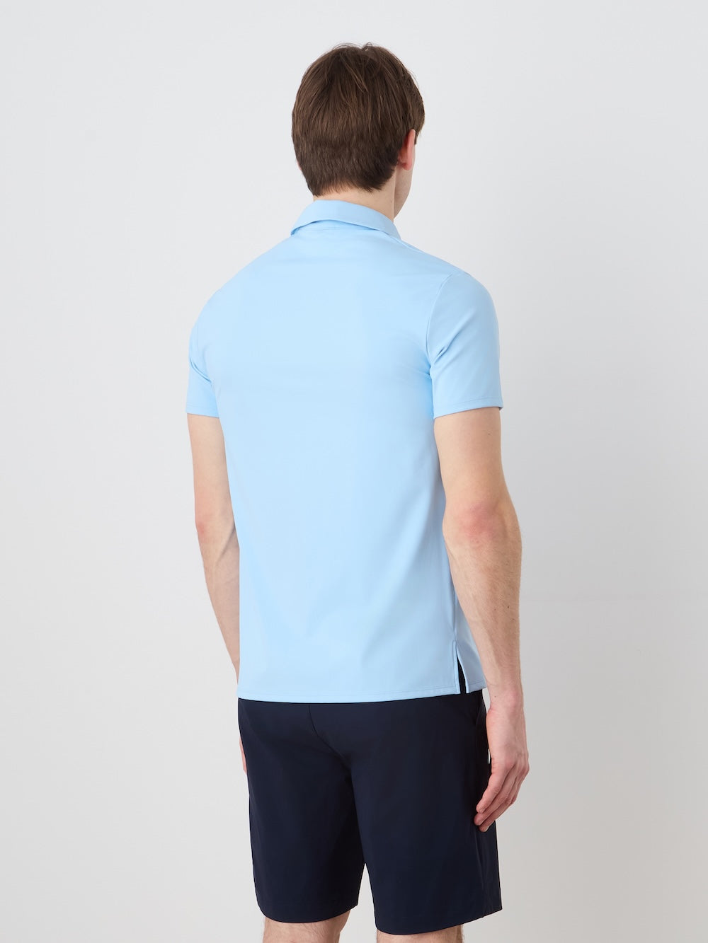 A person with short brown hair is facing away from the camera, wearing a light blue We Norwegians Honor Zipped Polo Men and dark navy shorts against a plain white background.