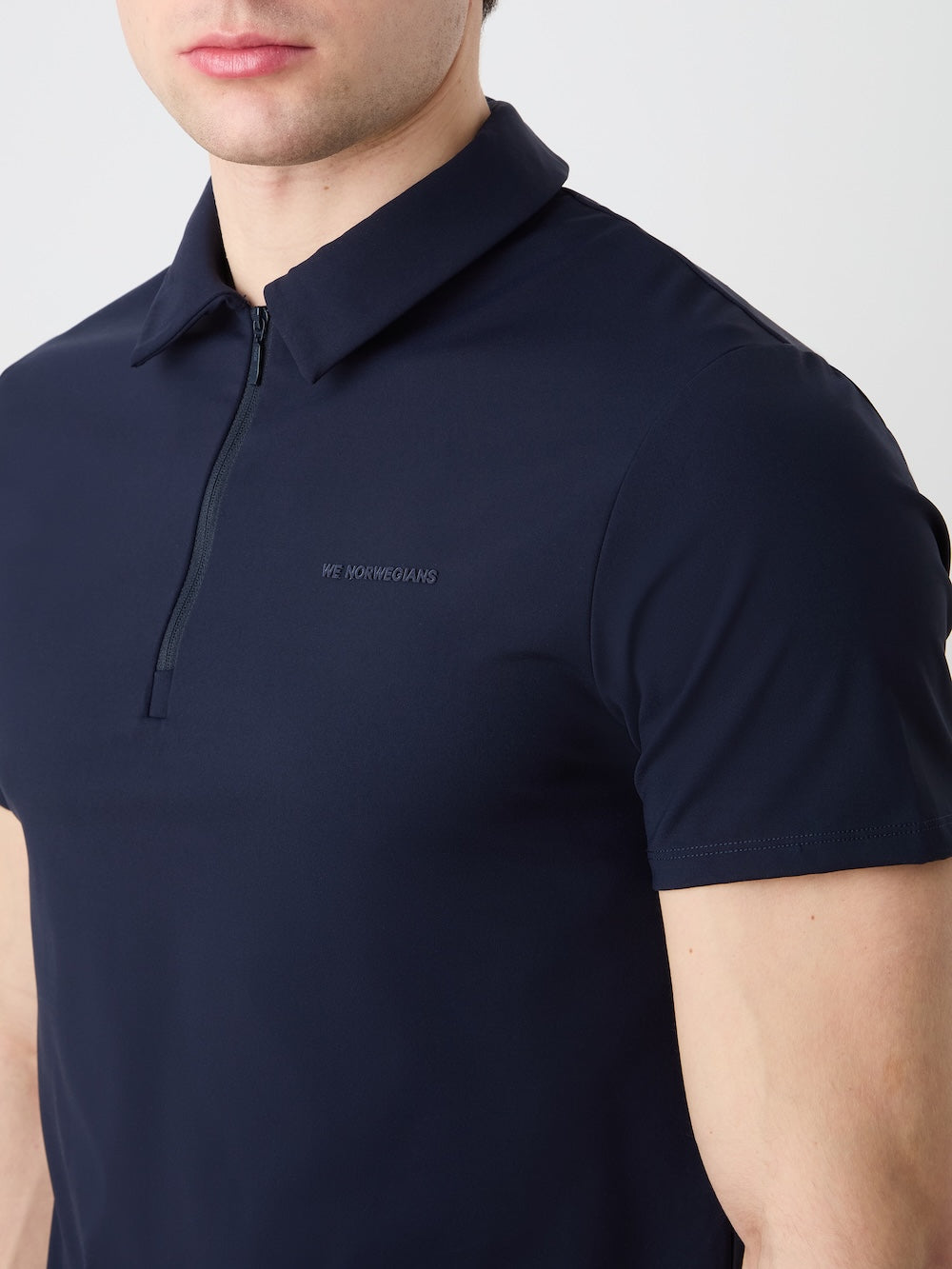 A person wears the We Norwegians Honor Zipped Polo Men in dark navy, featuring a zip-up collar and subtle chest branding. Made of high-performance fabric, only their lower face and upper torso are visible.