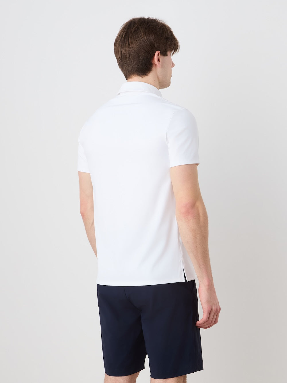A light-skinned man with brown hair, facing away from the camera, wears a white We Norwegians Honor Zipped Polo Men and navy shorts against a plain light background.