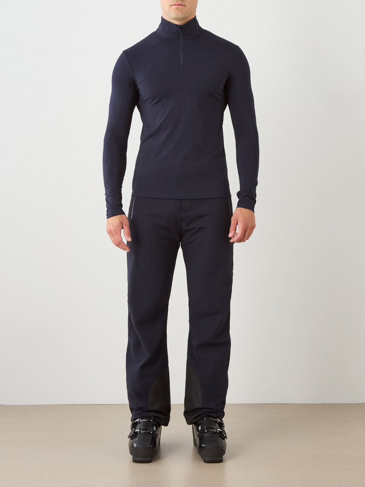 A person stands facing forward wearing the We Norwegians Honor Zipped Long Sleeve Men, paired with matching black pants and black ski boots, against a plain light background.