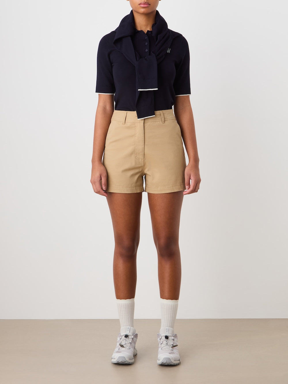 A person stands against a plain background wearing Sol Shorts Women in beige by We Norwegians, a black collared shirt, a matching sweater over their shoulders, white socks, and gray sneakers. Their arms rest naturally by their sides.