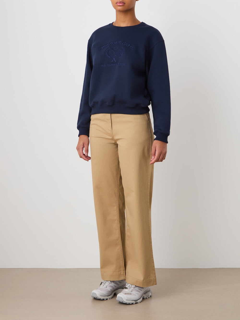 A person stands indoors against a plain white background, partially out of frame, wearing We Norwegians' Sol Pant Women in a tan cotton blend with a navy sweatshirt and gray sneakers.