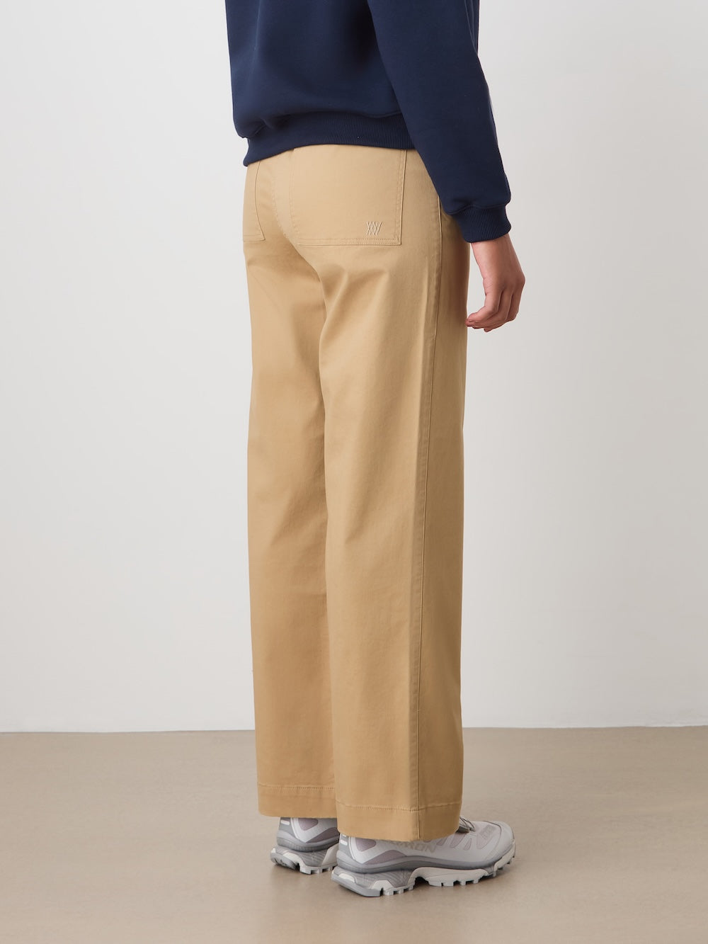 A person is seen from the back and side indoors, wearing We Norwegians Sol Pant Women—high-waisted, wide-legged beige pants—styled with a navy sweatshirt and white sneakers. The person's face is not visible.