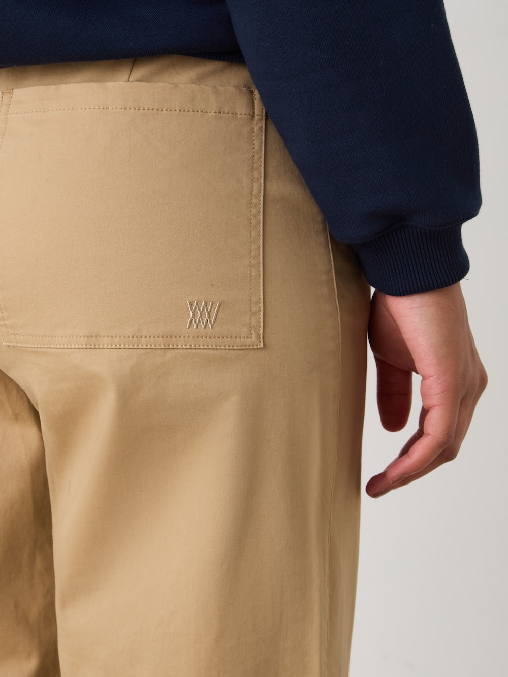 A person wears We Norwegians Sol Pant Women in high-waisted tan, featuring a visible back pocket, paired with a navy blue long-sleeve shirt. The photo highlights the pocket area and part of the right hand.