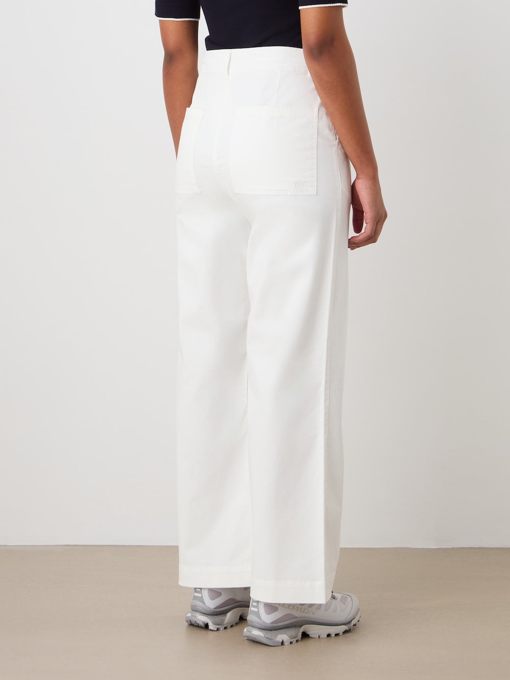 A person stands facing away, wearing We Norwegians' Sol Pant Women—high-waisted, wide-leg white sailor trousers—in a cotton blend, paired with a black short-sleeve top with white trim and light gray sneakers against a plain background.