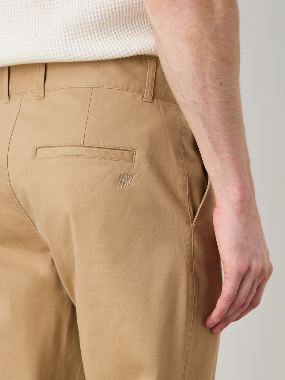 A man wears We Norwegians Sol Pant Men—tailored tan chinos in cotton stretch—and a white textured shirt, shown waist down and side-on, with his right hand beside his pocket.