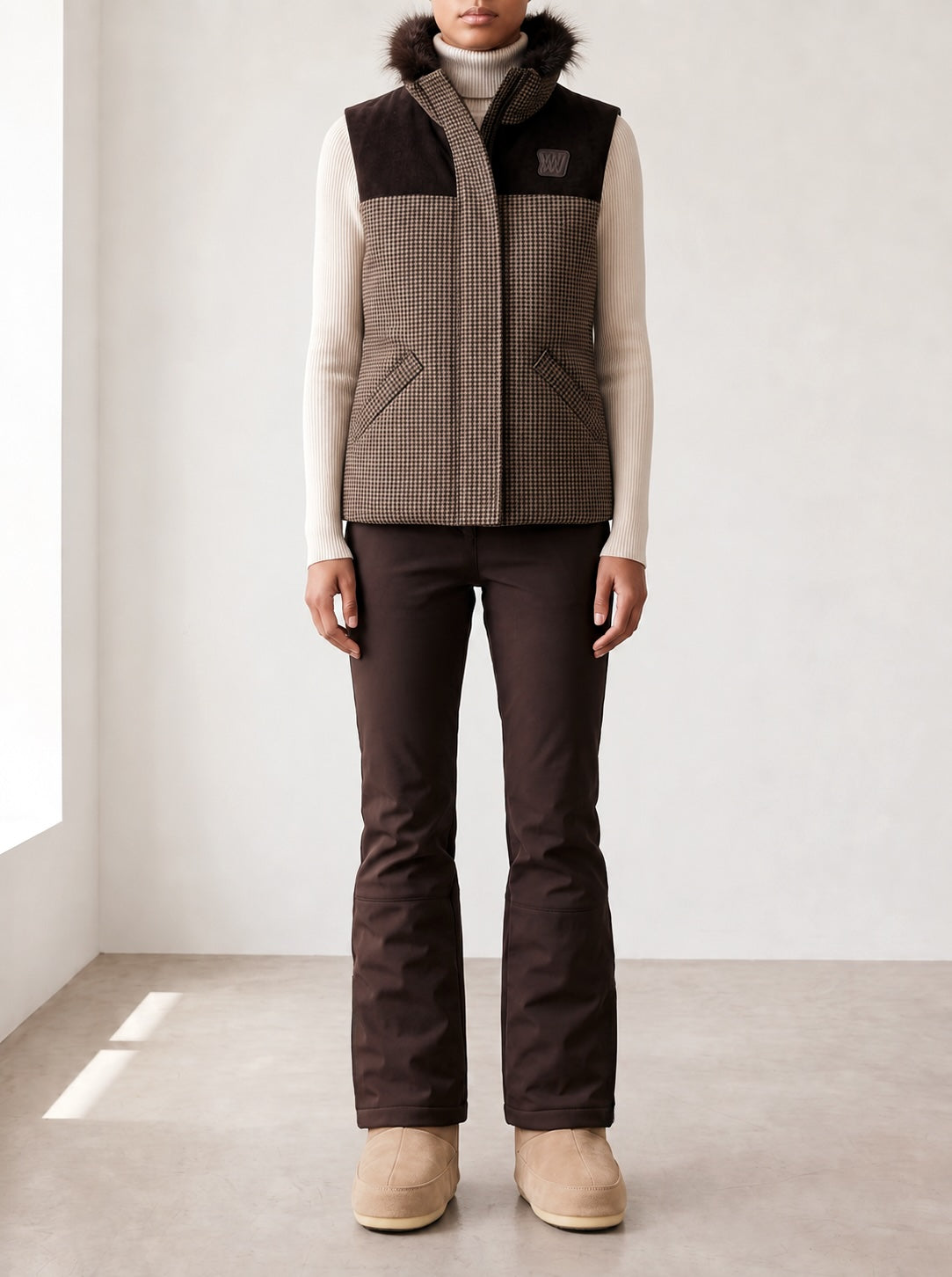 A woman models the We Norwegians Glenshee Vest, featuring a houndstooth pattern and faux fur collar, layered over a cream turtleneck sweater with dark brown pants and tan boots—an outfit that channels classic English countryside style against a soft, neutral backdrop.