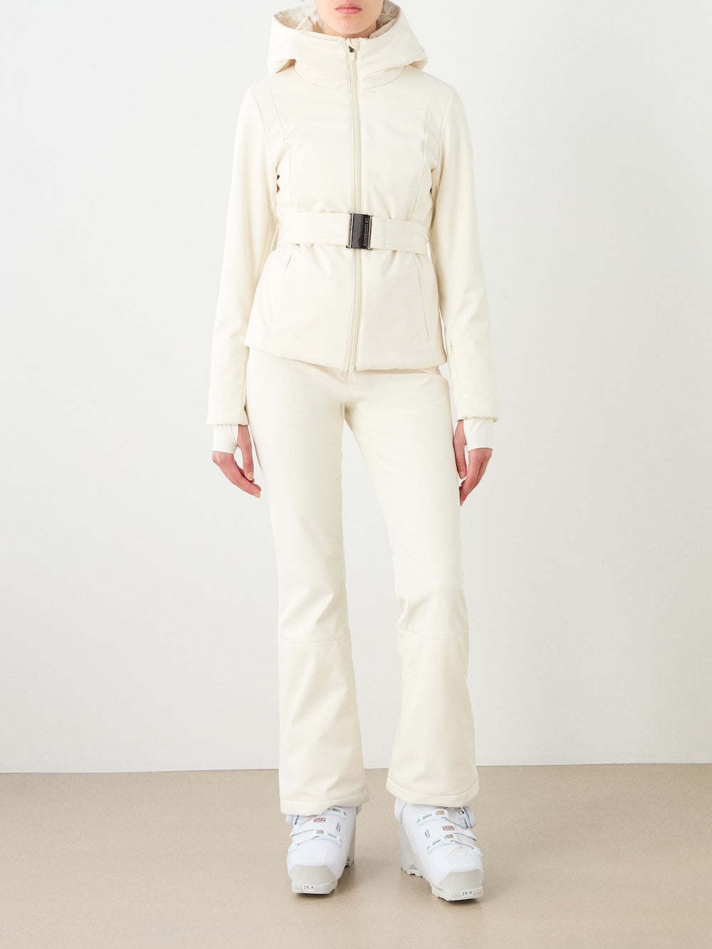 A woman wears the Tyin Tailored Ski Jacket Women by We Norwegians, styled with fitted pants and white ski boots, standing against a plain light background.