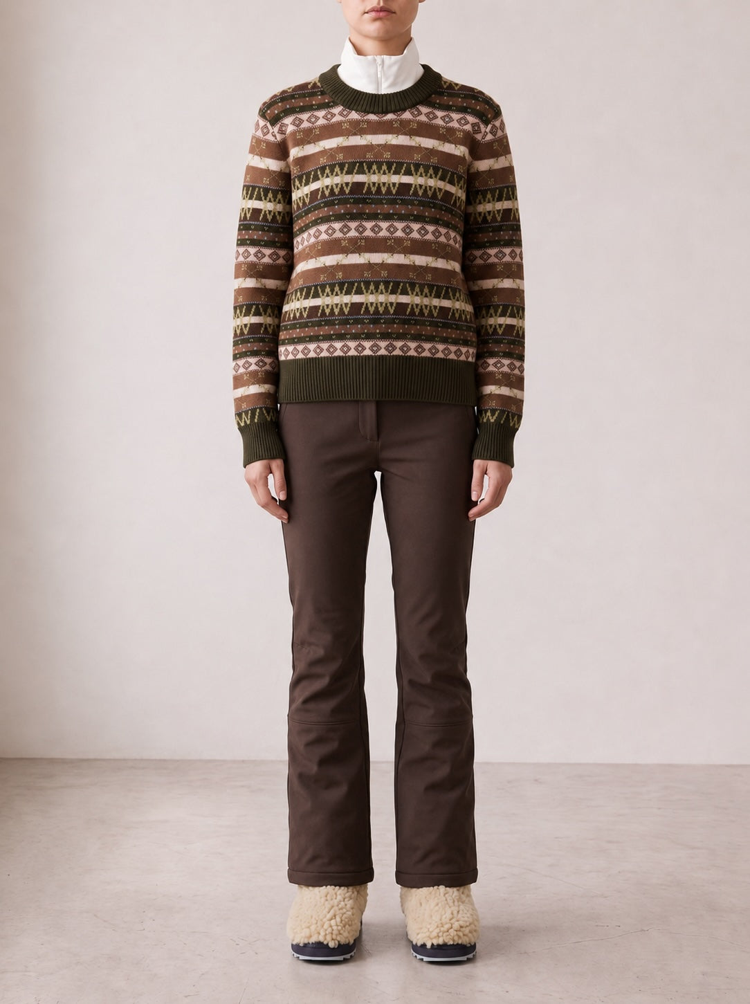 A person stands against a light wall, wearing the We Norwegians Skye Sweater Women—a Fair Isle knit merino wool sweater in earth tones—layered over a white turtleneck, paired with brown pants and cream-colored fuzzy boots. Their hands rest by their sides.