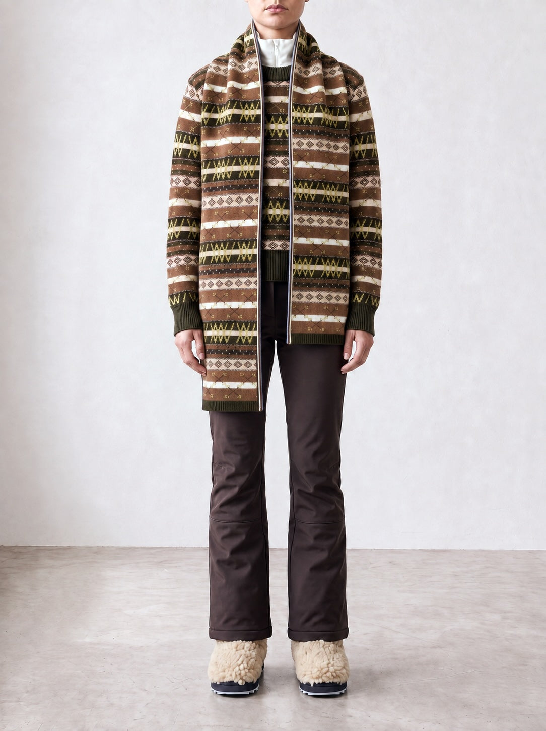 A person stands indoors against a plain light-colored wall and floor, wearing a patterned brown and green knit jacket, white turtleneck, dark brown pants, beige fuzzy boots, and accessorizing with the Skye Scarf by We Norwegians.