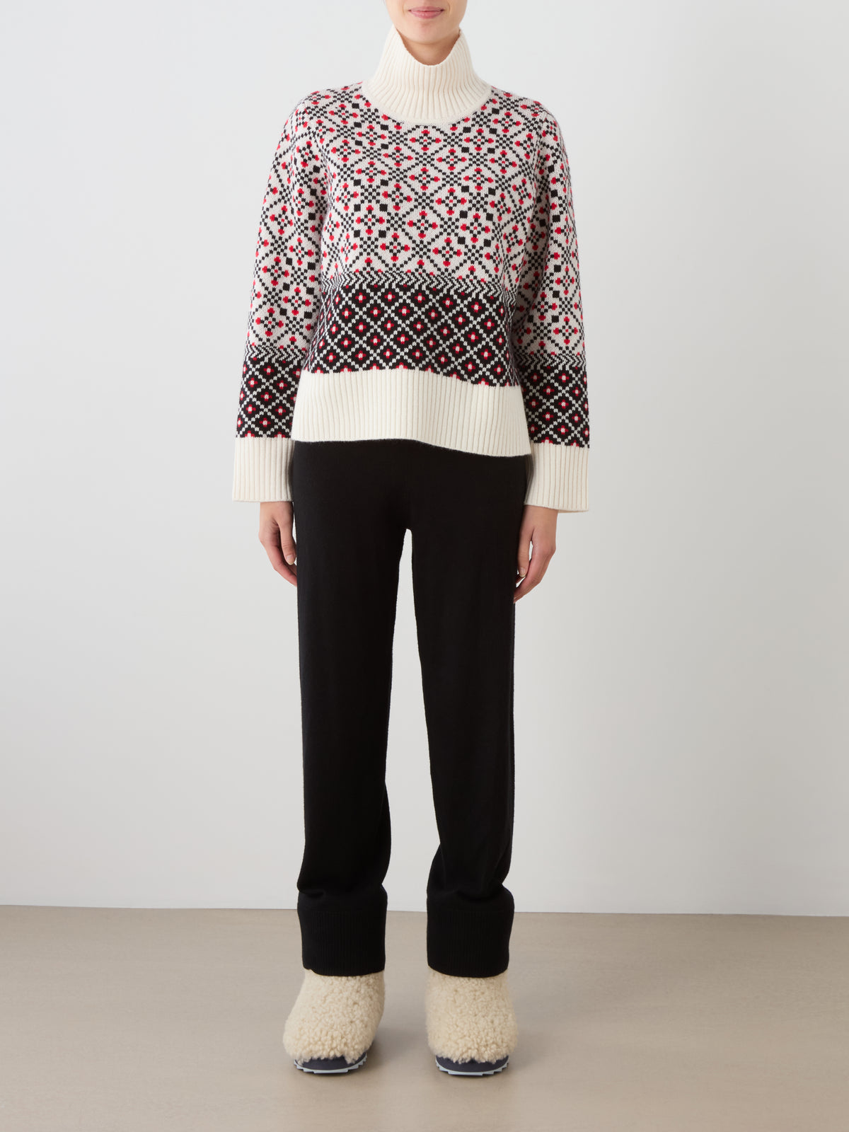 A person wearing the We Norwegians Telemark Sweater Women—a cream merino wool turtleneck featuring a red and black geometric Norwegian knit pattern—paired with black pants and cream fuzzy slippers, stands against a plain background, embodying a true winter essential.