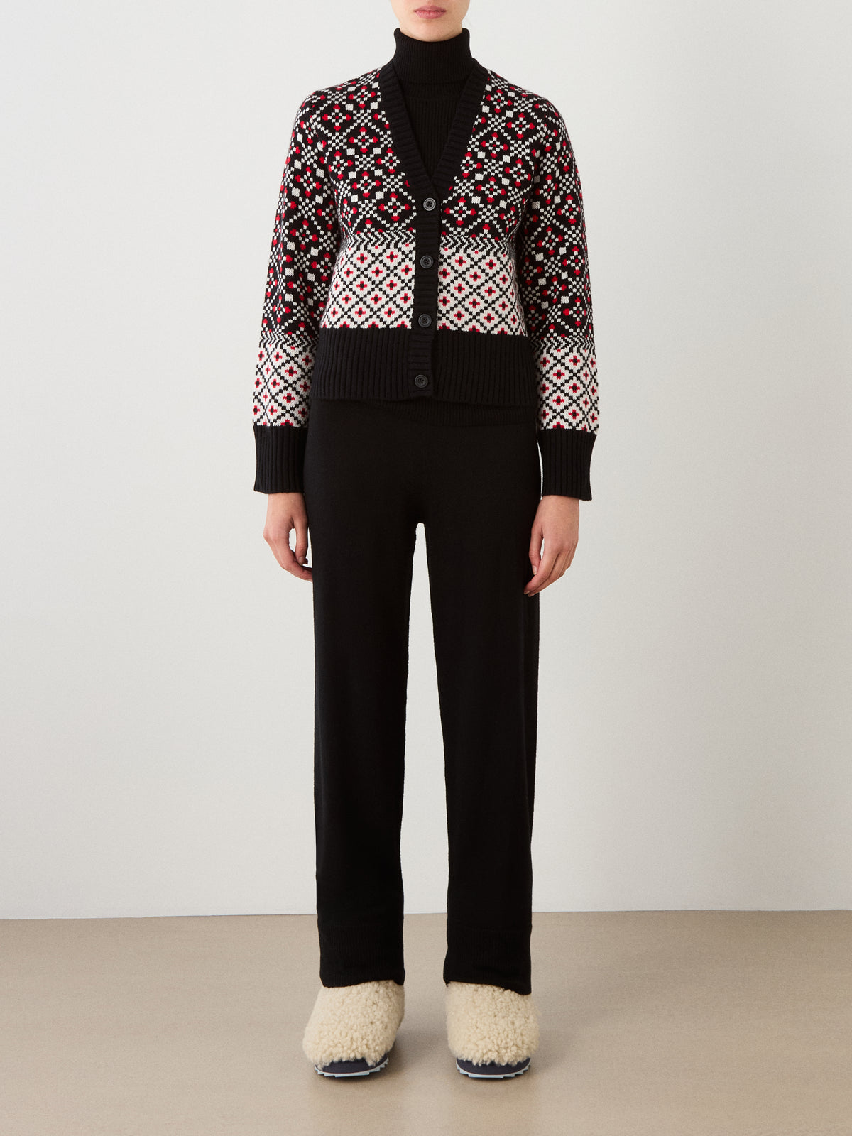A person wearing a black turtleneck, the We Norwegians Telemark Cardigan Women featuring red, white, and black Nordic-inspired designs, black pants, and cream-colored fuzzy slippers stands against a plain background.