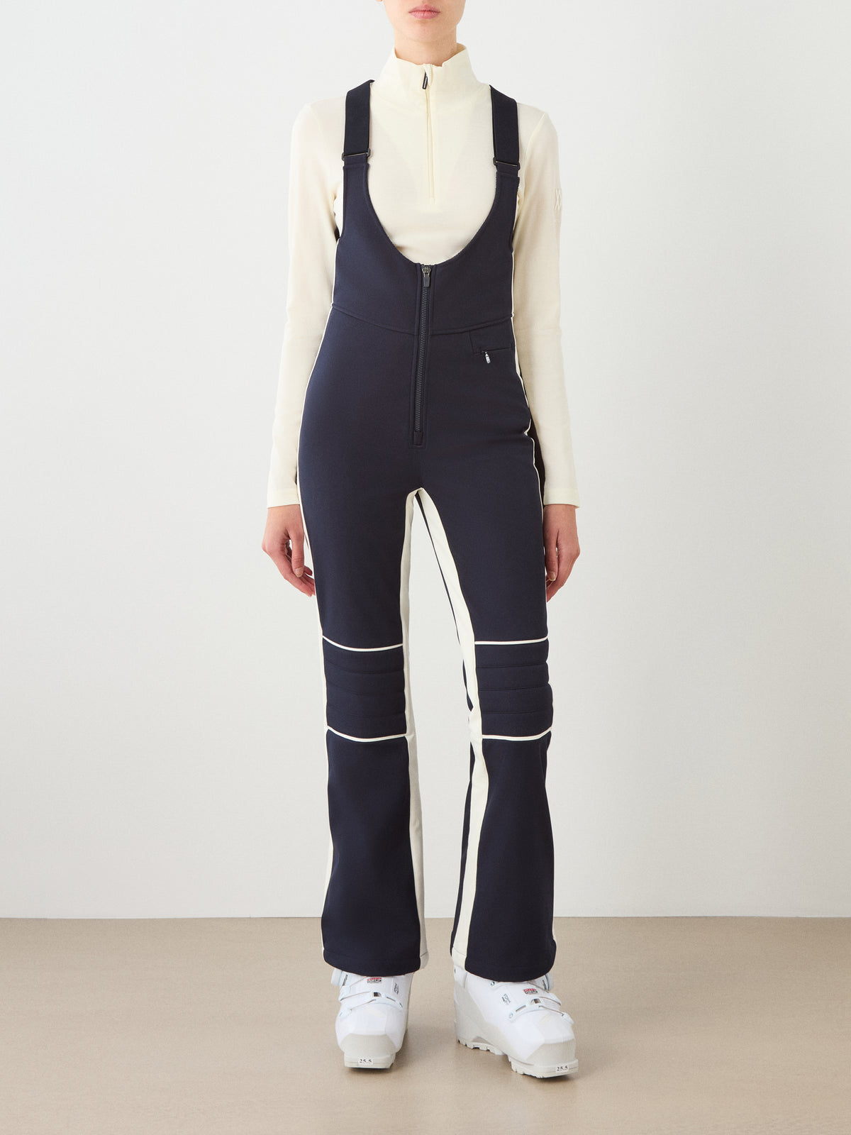 A person models the We Norwegians Solo Bib Pant Women in navy blue with white trim, paired with a cream zip-up top and white ski boots, embodying vintage alpine style against a plain light background.