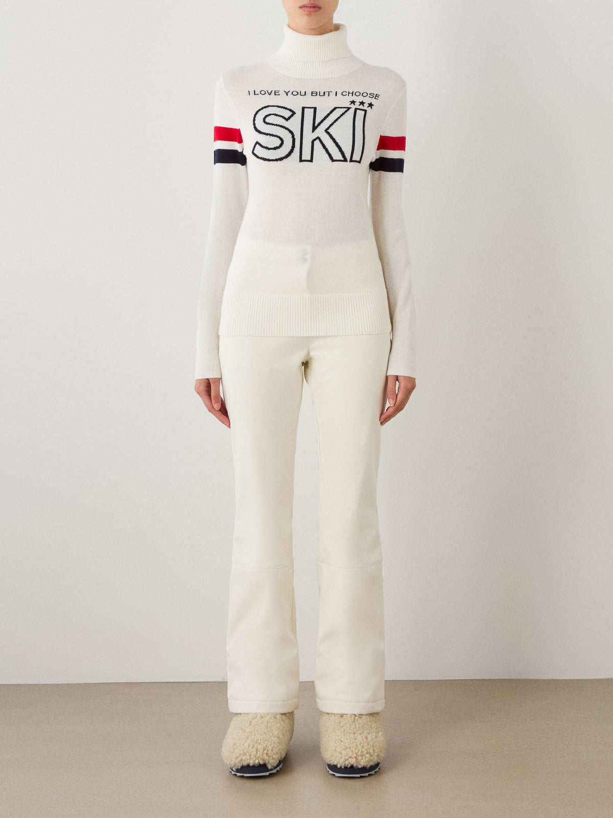 A person stands against a plain wall wearing the We Norwegians Bibi Fitted Sweater Women, a white top with “SKI” text and red and black striped sleeves, paired with white fitted pants and beige fuzzy boots for a retro ski-inspired look.