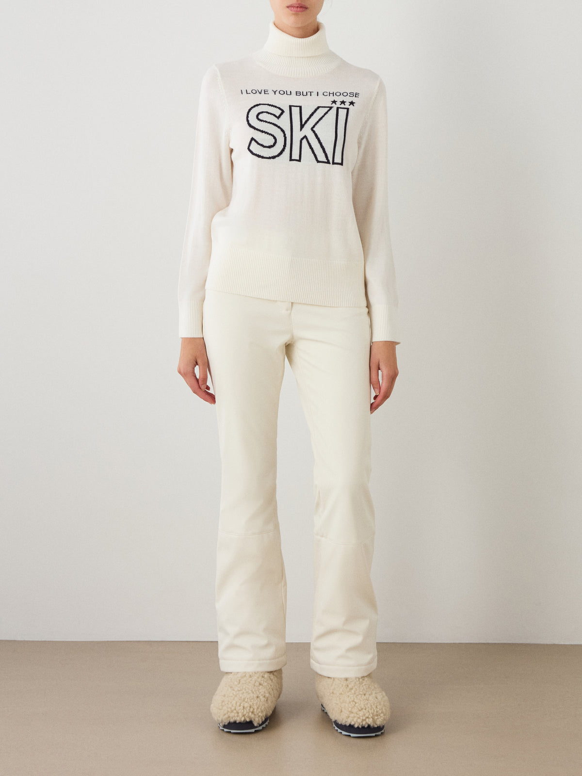 A person stands against a white wall wearing an all-white outfit: the We Norwegians Bibi Sweater Women featuring a retro ski fashion-inspired design with “I LOVE YOU BUT I CHOOSE SKI” printed on it, paired with fitted pants and fluffy white boots.