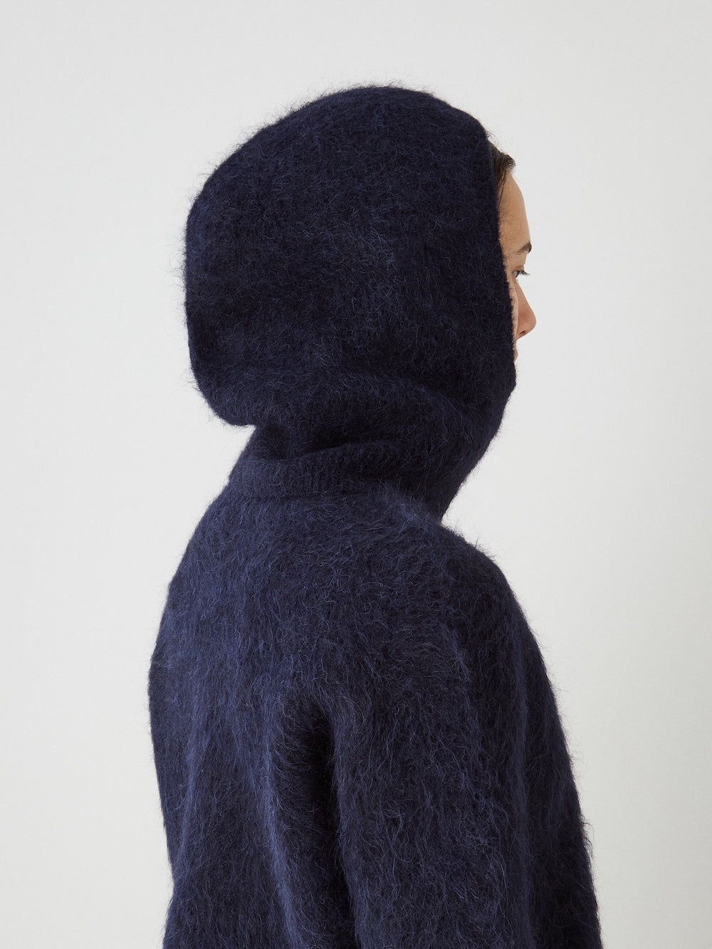 Person in profile wearing the We Norwegians Myrull Balaclava, hood up, resembling a cozy alpaca knit, against a plain light background. Only part of their face is visible.