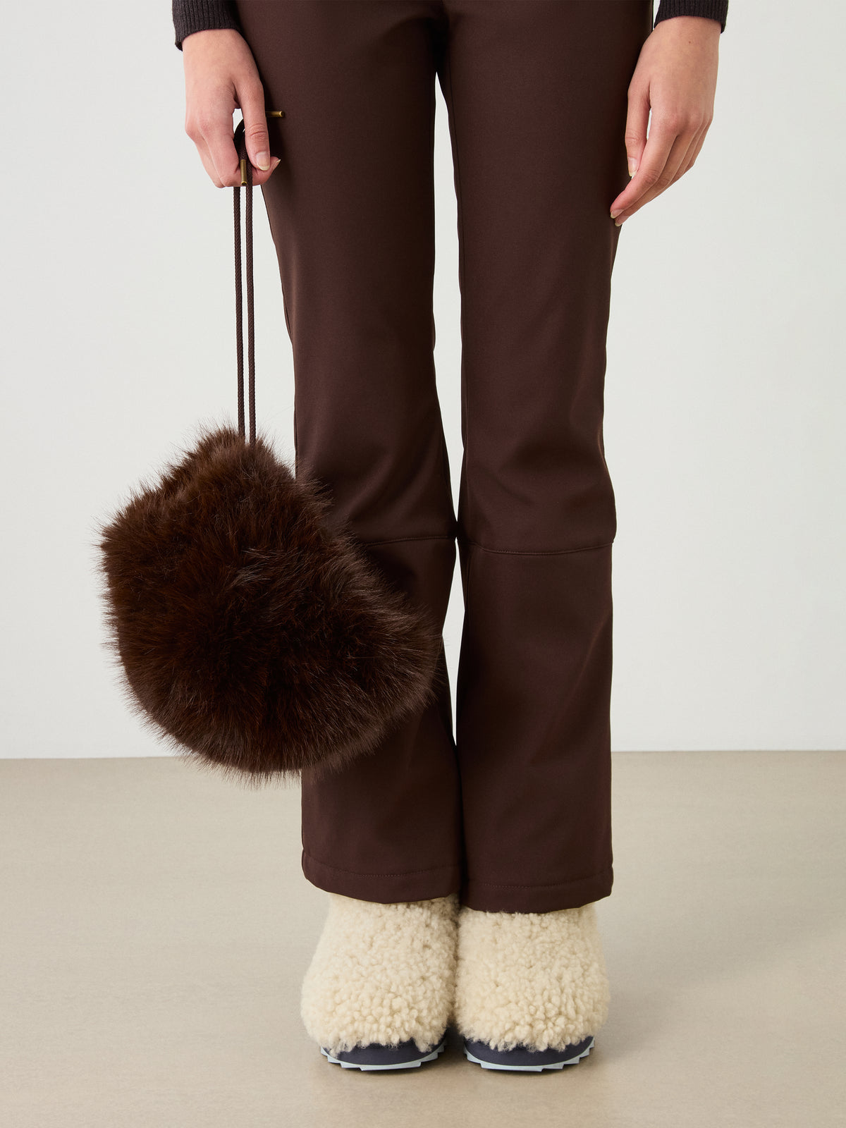 A person in brown pants, the We Norwegians Dovre Bonnet, and cream fuzzy slippers holds a round brown furry handbag by a thin strap, standing on a light floor against a plain background.