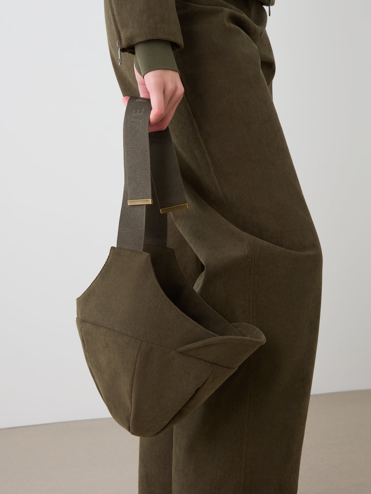 Wearing olive green baby corduroy, a person holds a matching wide-strapped handbag against a light background—an ideal setting to showcase the Britt Bonnet by We Norwegians.