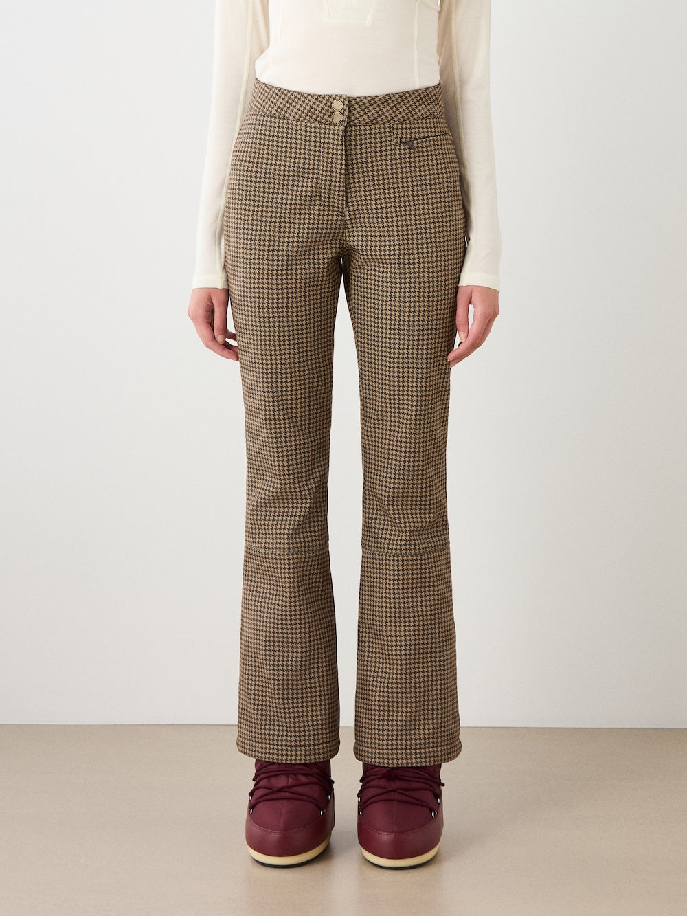 Wearing Glenshee Ski Pant Women by We Norwegians in brown and beige houndstooth, paired with a white long-sleeve top and maroon boots; shown from shoulders down against a neutral background.