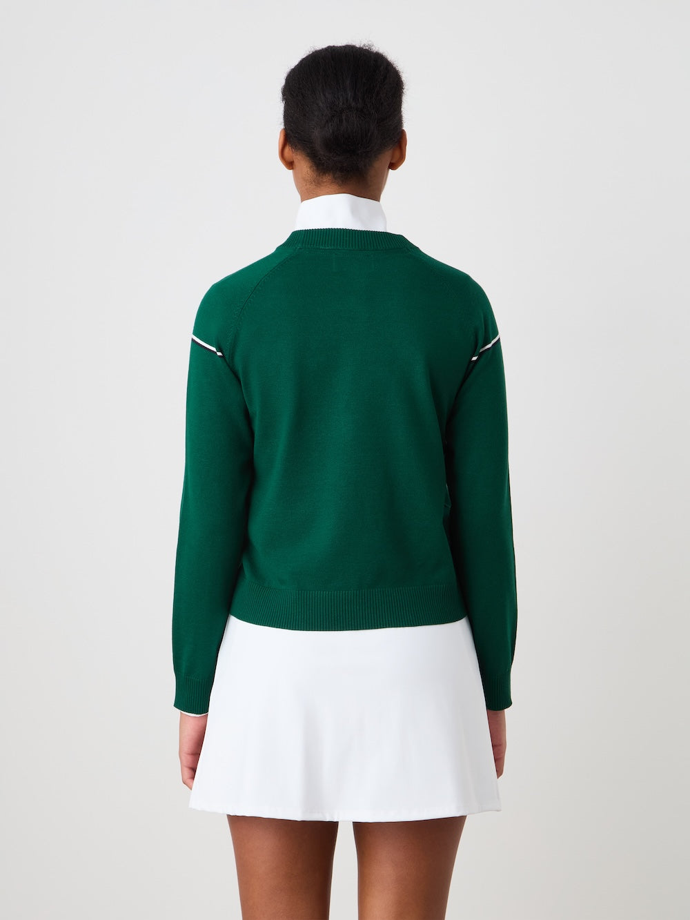 A woman faces away from the camera in a We Norwegians Grand Slam Sweater, layered over a white collared shirt and skirt, showcasing vintage athletic style against a simple light backdrop.