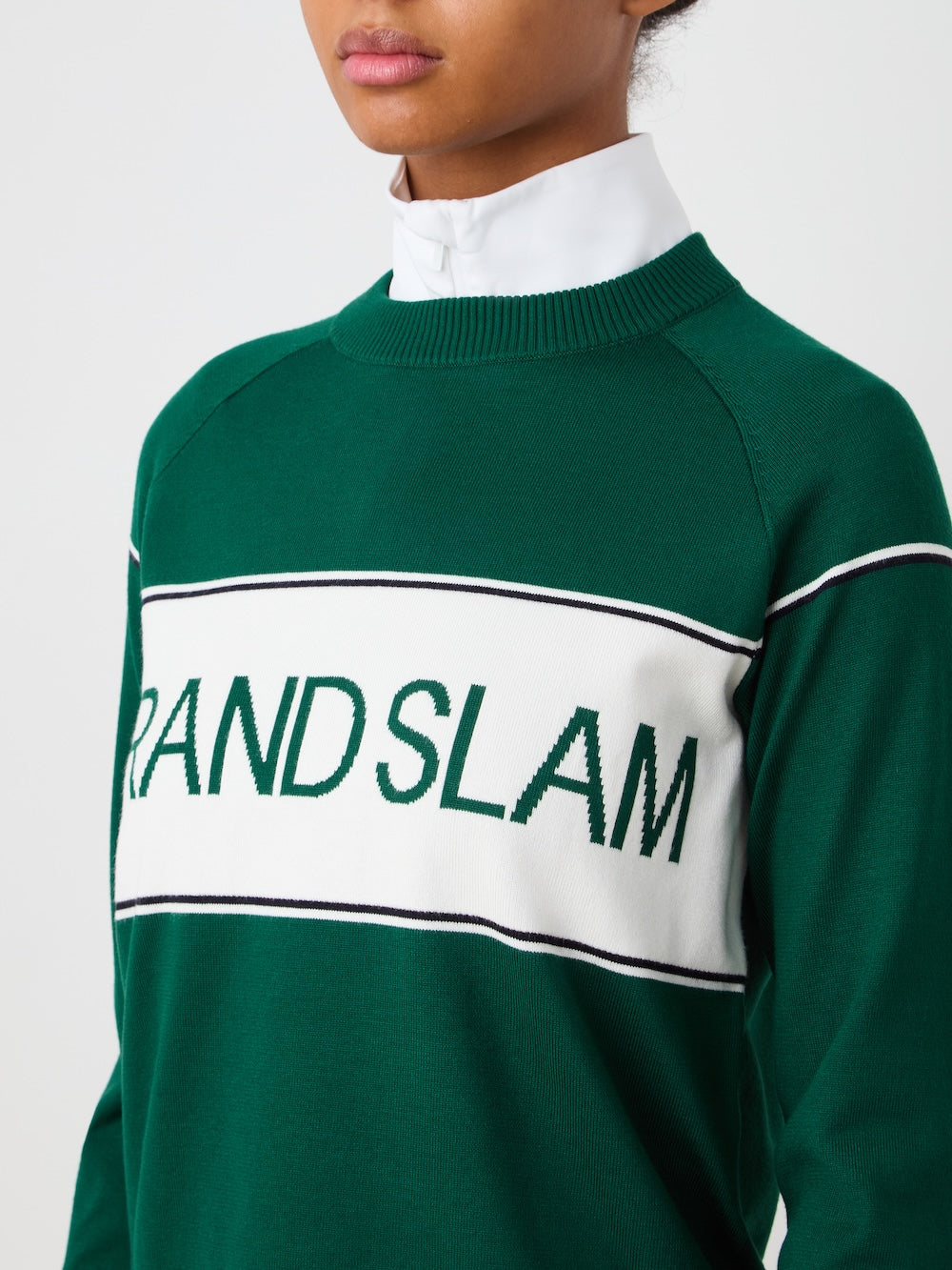 A woman wears the We Norwegians Grand Slam Sweater—a green vintage athletic-style knit with a white chest panel and "GRAND SLAM" in green—layered over a white collared shirt, against a plain background with her face partially visible.