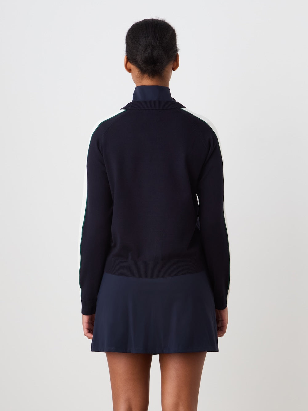 A woman with dark hair in a low bun is seen from behind, wearing the We Norwegians Grand Slam Sweater Women in navy with striped sleeves and a matching skirt, channeling vintage athletic style against a plain white background.