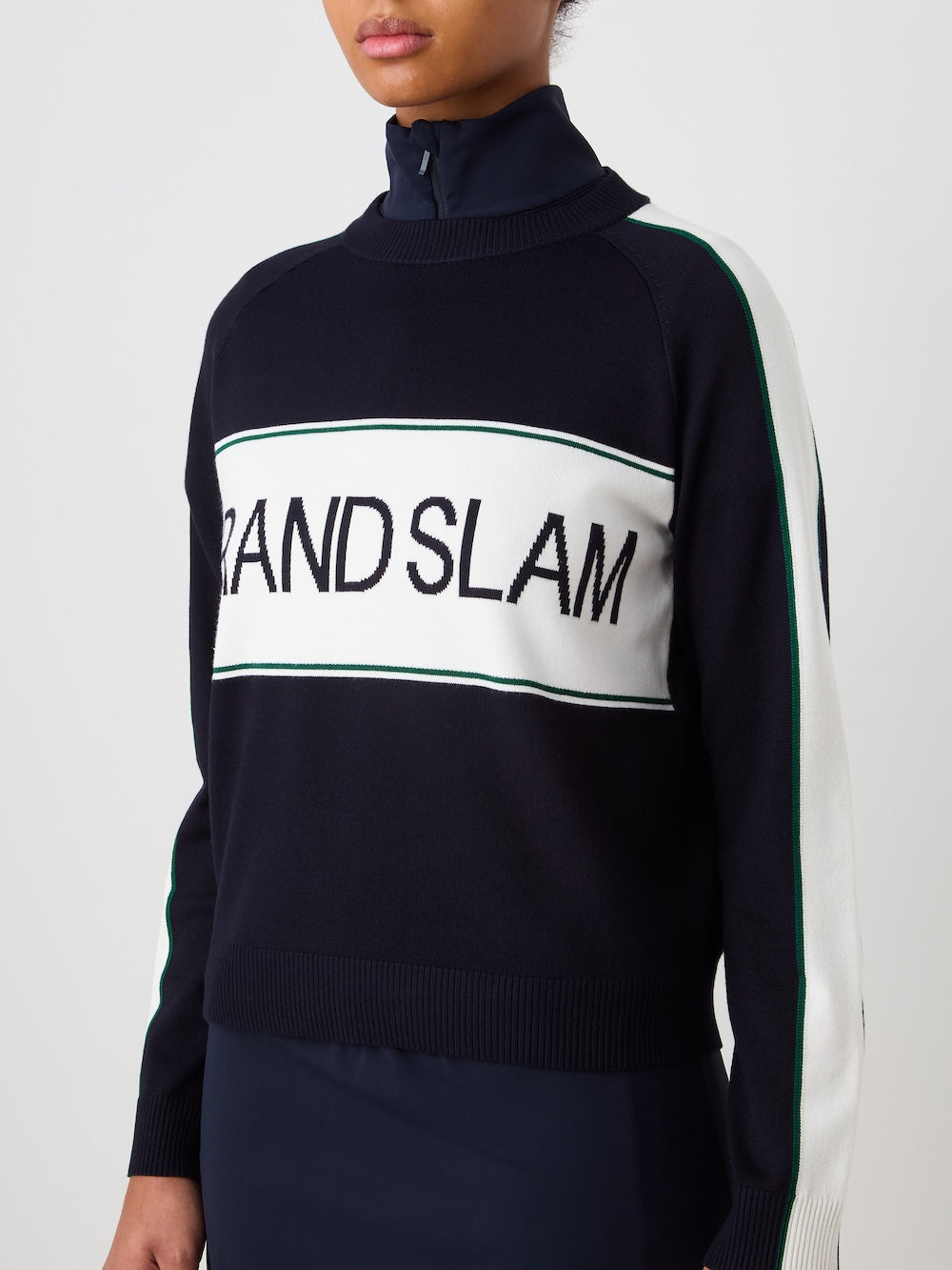 A woman wears the We Norwegians Grand Slam Sweater—navy blue with white and green stripes and "GRAND SLAM" across the chest—layered over a dark collared shirt, shown from the chest up.