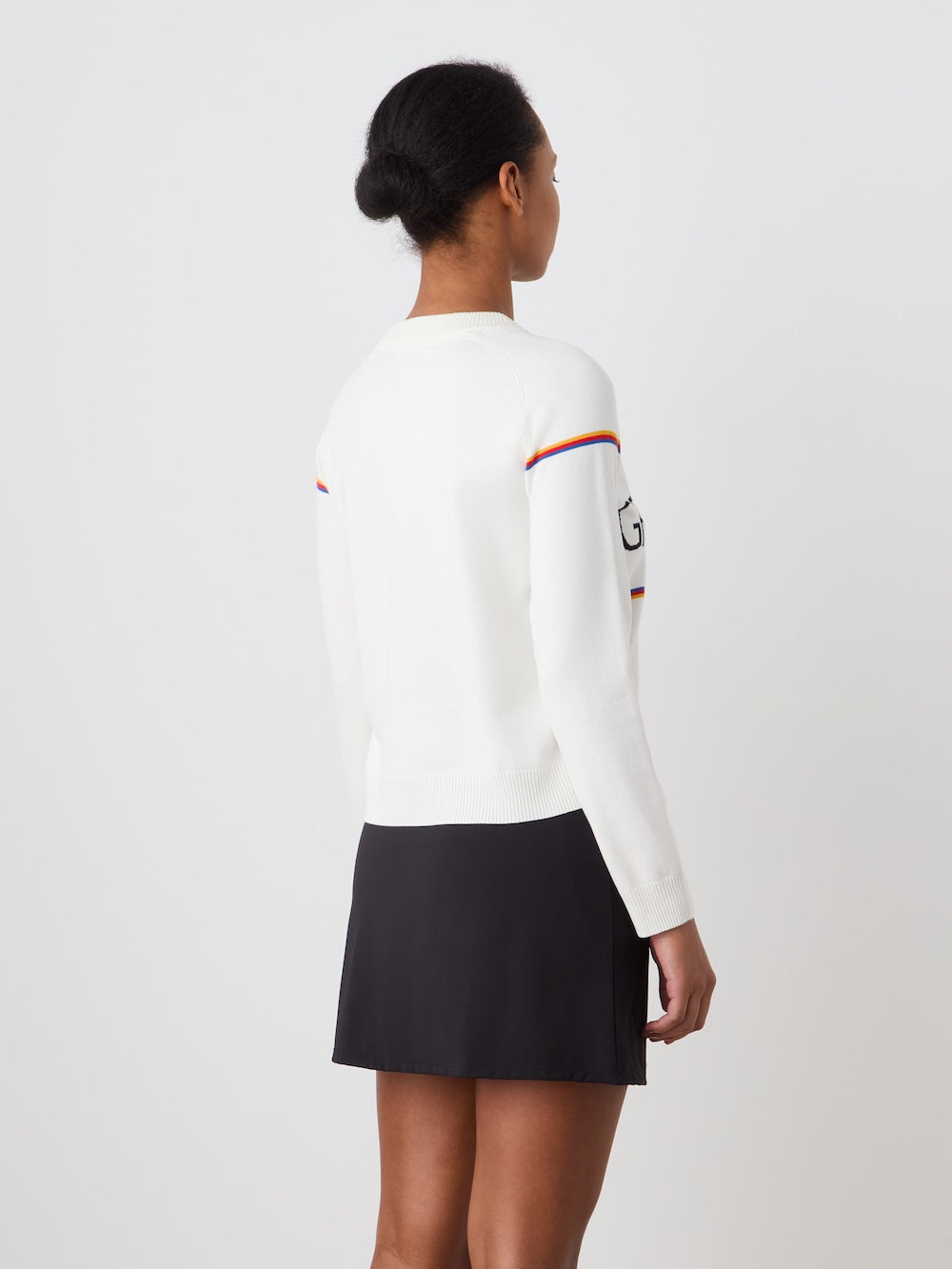 A woman with her hair in a neat bun models the We Norwegians Grand Slam Sweater Women—white with colored sleeve stripes—and a short black skirt, standing against a plain white background, facing away from the camera.